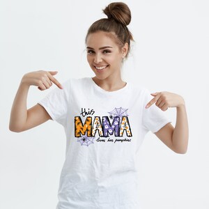 Pode incluir: Camiseta branca com um gr&aacute;fico tem&aacute;tico de Halloween que diz "This Mama loves her pumpkins". O gr&aacute;fico apresenta uma "Mama" roxa e laranja com teias de aranha e fantasmas.