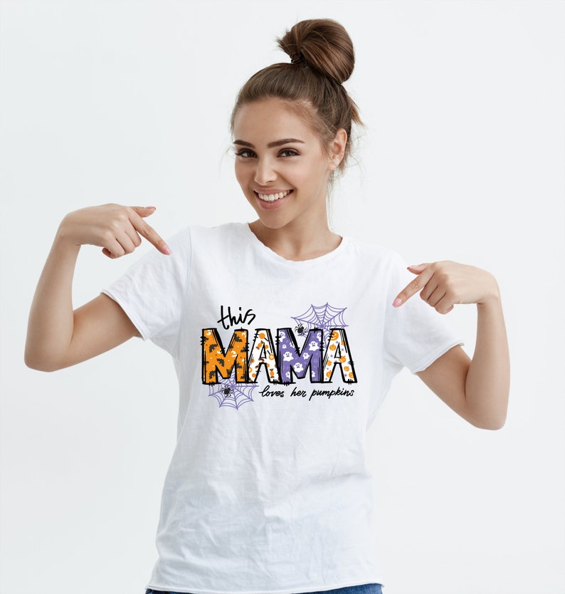 Pode incluir: Camiseta branca com um gr&aacute;fico tem&aacute;tico de Halloween que diz "This Mama loves her pumpkins". O gr&aacute;fico apresenta uma "Mama" roxa e laranja com teias de aranha e fantasmas.