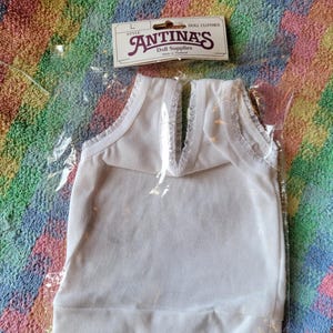 May include: White sleeveless doll top with a lace trim around the neckline. The top is packaged in a clear plastic bag with a label that reads "Antina's Doll Supplies" and "Doll Clothes".