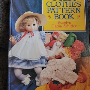 May include: A vintage book titled "Dolls' Clothes Pattern Book" with a doll wearing a white dress and straw hat. The cover is blue with a yellow box containing the title and author's name. Various doll clothes and sewing notions are visible.