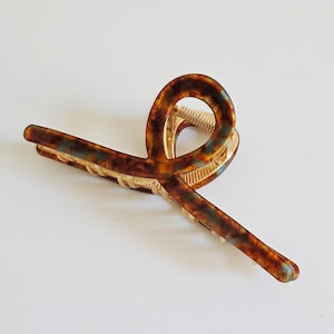 May include: A tortoiseshell hair claw clip with a gold-colored metal clasp. The clip features a curved design with a marbled pattern in shades of brown, amber, and hints of blue-green. This accessory is suitable for various hairstyles.