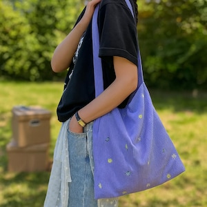 Puede incluir: Un bolso tote de pana lavanda con una correa ancha para el hombro. El bolso presenta diseños florales amarillos y verdes bordados. La persona de la imagen lleva una camiseta negra y vaqueros azules.