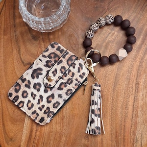 May include: A leopard print cardholder with a gold zipper and snap closure. A matching wristlet keychain with a tassel and dark brown beads is attached. The keychain has a gold clasp and a wooden geometric bead.