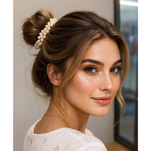 May include: A woman with brown hair styled in a bun, adorned with a double-strand pearl hair clip. She wears a white top and a delicate gold necklace. The image is a close-up, showcasing her makeup and a soft, natural glow.