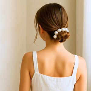 May include: A woman's hair is styled in a bun, adorned with a pearl hair accessory. The hair is a dark brown color, and the woman is wearing a white tank top. The pearls are large and round, adding a touch of elegance.