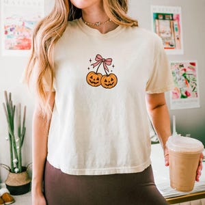 以下が含まれることがあります： ハロウィーンをテーマにしたデザインのクリーム色のTシャツ。デザインには、笑顔の顔をした2つのオレンジ色のジャック・オー・ランタンが、茎とピンクのリボンでつながっています。Tシャツは半袖で、クルーネックです。