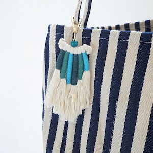 May include: A close-up of a blue and white striped tote bag with a macrame tassel keychain. The keychain features a gold clasp, a turquoise bead, and a tassel with teal, blue, and cream-colored threads. The tote bag has a striped pattern.