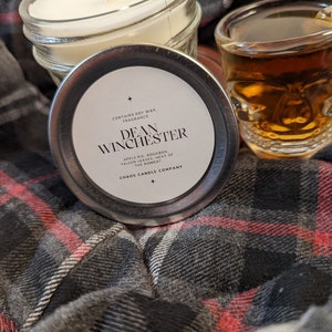 May include: A candle in a glass jar with a silver lid. The lid has a label that says "CONTAINS SOY WAX. FRAGRANCE DEAN WINCHESTER APPLE PIE, BOURBON, FALLEN LEAVES, HEAT OF THE MOMENT CHAOS CANDLE COMPANY".