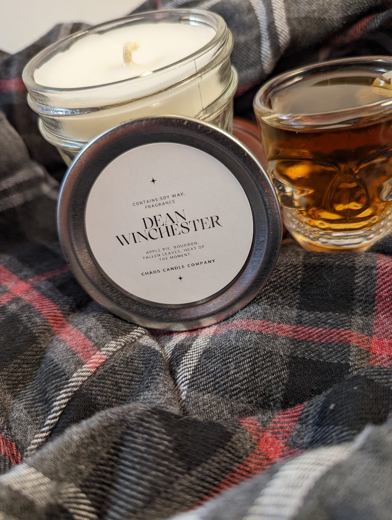 May include: A candle in a glass jar with a silver lid. The lid has a label that says "CONTAINS SOY WAX. FRAGRANCE DEAN WINCHESTER APPLE PIE, BOURBON, FALLEN LEAVES, HEAT OF THE MOMENT CHAOS CANDLE COMPANY".
