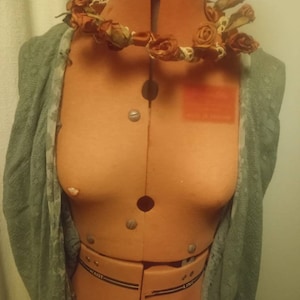 May include: An orange dressmaker's mannequin with a floral garland around the neck and a grey shawl draped over the shoulders. The garland features small, dried orange roses. The mannequin has a waist and bust line marked with black lines and metal studs.