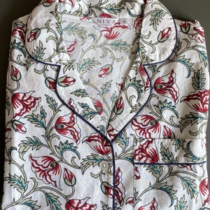 May include: White cotton pajama top with a floral print featuring red, green, and blue colors. The top has a collar, button closure, and a pocket on the left side. The label on the top reads "NIYA S".