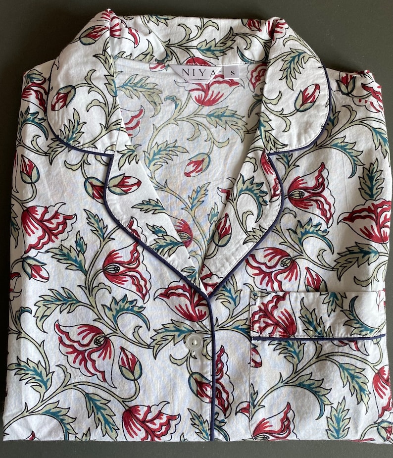 May include: White cotton pajama top with a floral print featuring red, green, and blue colors. The top has a collar, button closure, and a pocket on the left side. The label on the top reads "NIYA S".
