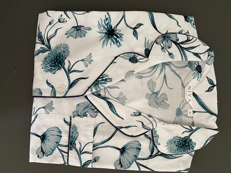 May include: White pajama top with a blue floral print and navy blue trim. The top has a button closure and a pocket on the left side. The brand name "NIYA" is printed on the tag.