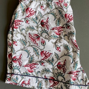 May include: White cotton pajama shorts with a floral print featuring red and green flowers. The shorts have a navy blue trim around the edges.