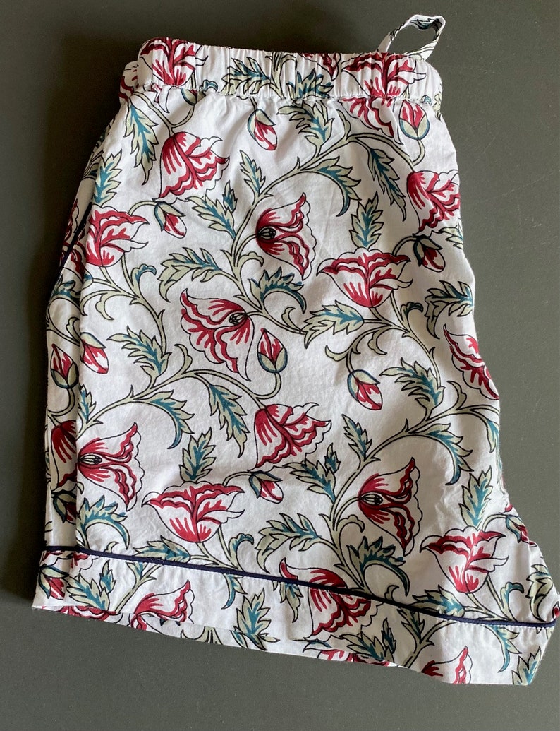 May include: White cotton pajama shorts with a floral print featuring red and green flowers. The shorts have a navy blue trim around the edges.