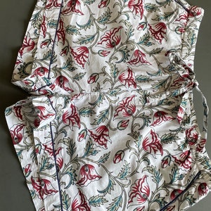 May include: White cotton shorts with a floral print featuring red and green flowers. The shorts have a navy blue trim and a drawstring waist.