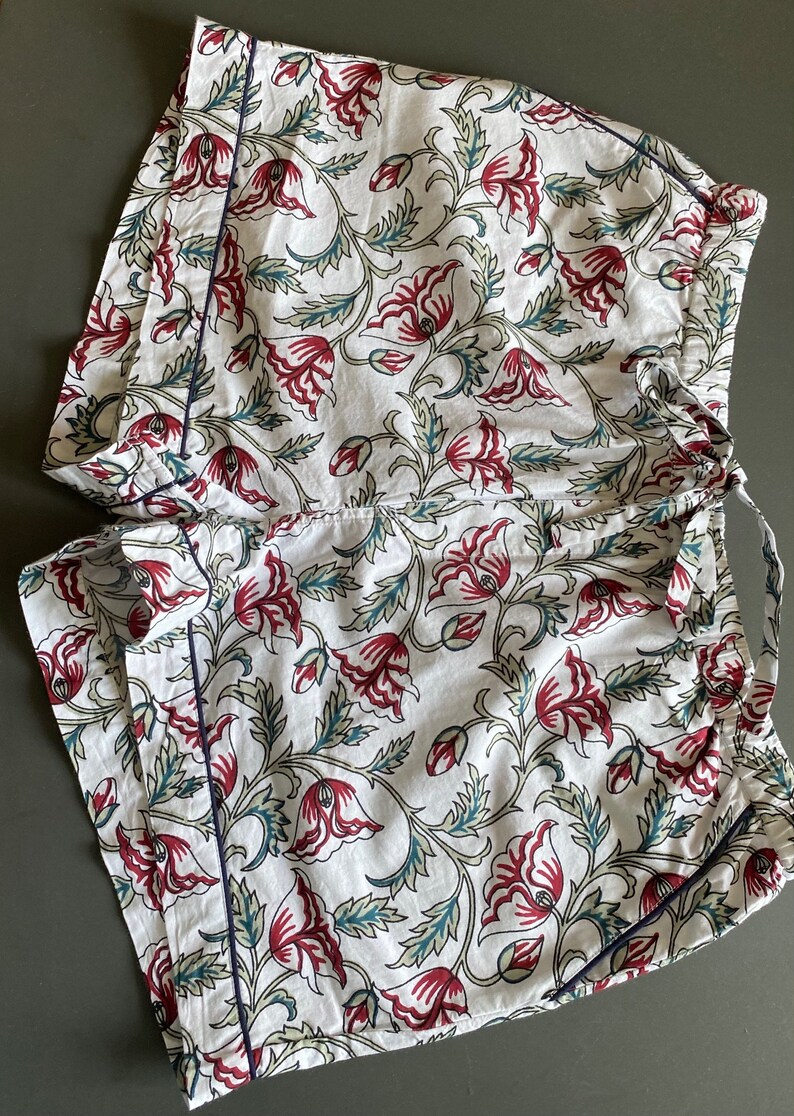 May include: White cotton shorts with a floral print featuring red and green flowers. The shorts have a navy blue trim and a drawstring waist.