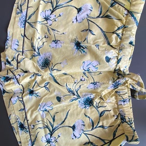 May include: Yellow cotton shorts with a floral print in shades of white and blue. The shorts have a black trim and a drawstring waist.