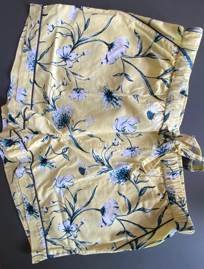 May include: Yellow cotton shorts with a floral print in shades of white and blue. The shorts have a black trim and a drawstring waist.