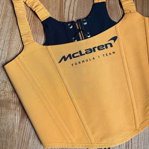 Può includere: Top a corsetto giallo senape con dettagli neri e il logo McLaren Formula 1 Team. Presenta spalline regolabili, allacciatura frontale e cuciture verticali. Il corsetto è realizzato in tessuto liscio.