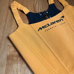 Może przedstawiać: Musztardowy gorset z czarnymi akcentami i logo McLaren Formula 1 Team. Top posiada zapięcie na zamek błyskawiczny i regulowane ramiączka. Gorset ma pionowe szwy i strukturalny design.