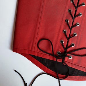Puede incluir: Un cors&eacute; rojo con cordones negros y ojales plateados. El cors&eacute; est&aacute; hecho de una tela lisa y brillante y tiene un dise&ntilde;o estructurado. Los cordones negros est&aacute;n atados en un patr&oacute;n entrecruzado y el cors&eacute; tiene un forro negro.