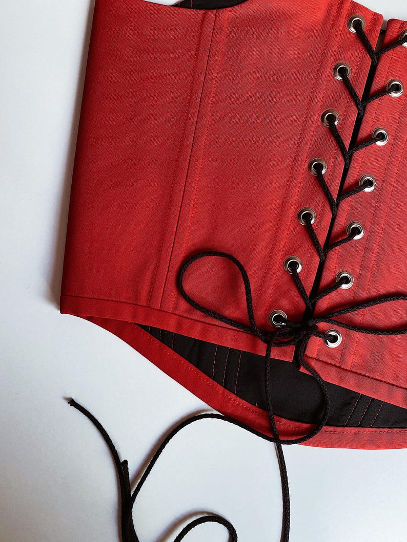 Puede incluir: Un cors&eacute; rojo con cordones negros y ojales plateados. El cors&eacute; est&aacute; hecho de una tela lisa y brillante y tiene un dise&ntilde;o estructurado. Los cordones negros est&aacute;n atados en un patr&oacute;n entrecruzado y el cors&eacute; tiene un forro negro.