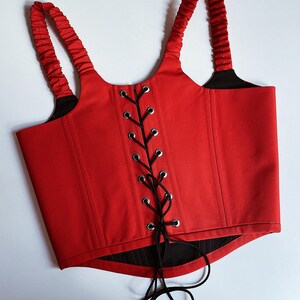 Puede incluir: Un cors&eacute; rojo con cordones y correas negras. El cors&eacute; presenta un dise&ntilde;o estructurado con un cierre central con cordones y ojales plateados. Las correas est&aacute;n fruncidas.