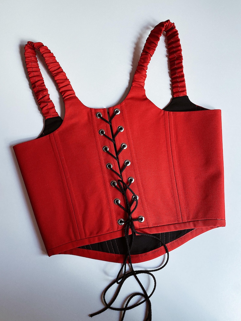 Puede incluir: Un cors&eacute; rojo con cordones y correas negras. El cors&eacute; presenta un dise&ntilde;o estructurado con un cierre central con cordones y ojales plateados. Las correas est&aacute;n fruncidas.