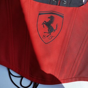 Puede incluir: Cors&eacute; rojo con el logo de Ferrari estampado en la parte delantera. El cors&eacute; tiene un forro negro y una espalda con cordones. El logo de Ferrari presenta un caballo encabritado negro sobre un escudo amarillo.