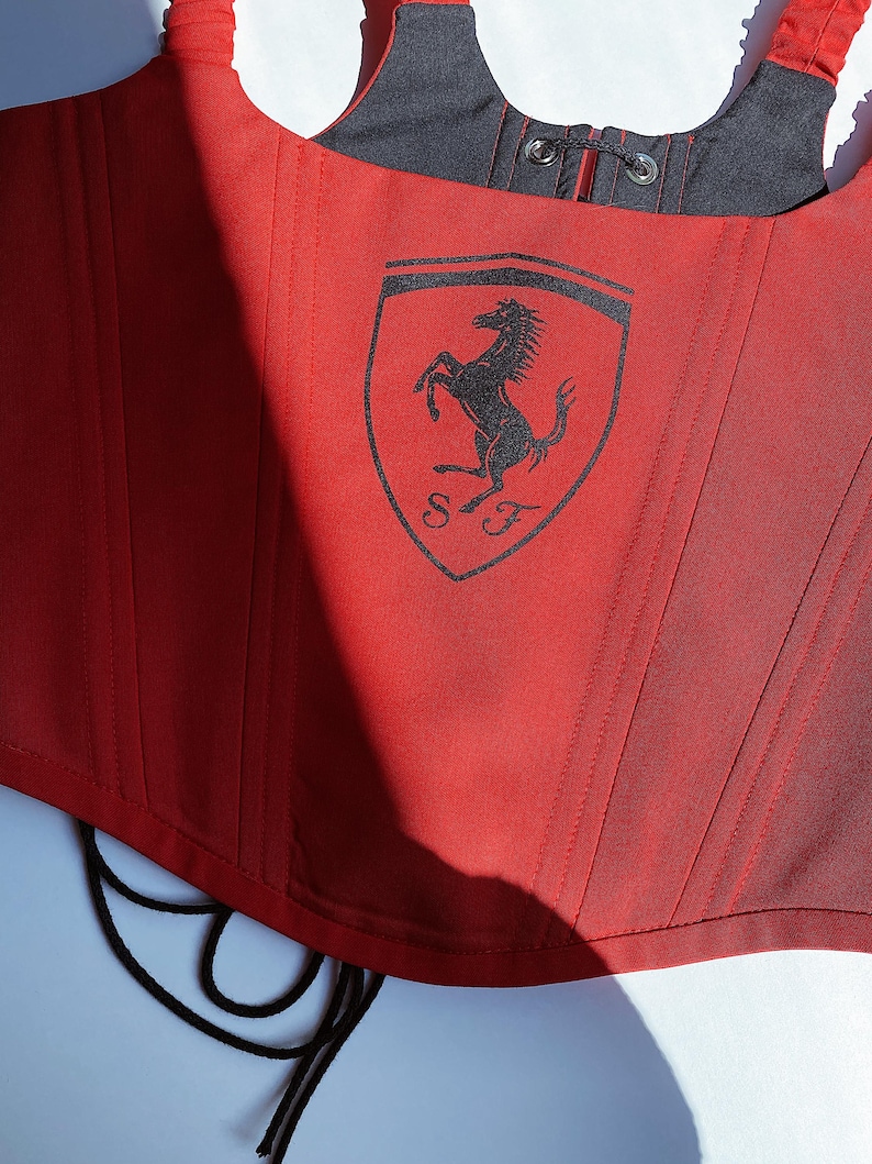 Puede incluir: Cors&eacute; rojo con el logo de Ferrari estampado en la parte delantera. El cors&eacute; tiene un forro negro y una espalda con cordones. El logo de Ferrari presenta un caballo encabritado negro sobre un escudo amarillo.