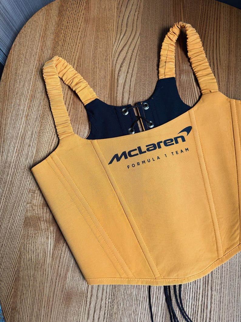 Może przedstawiać: Musztardowy gorset z czarnymi akcentami i logo McLaren Formula 1 Team. Top ma regulowane ramiączka, sznurowanie z przodu i pionowe szwy. Gorset jest wykonany z gładkiej tkaniny.
