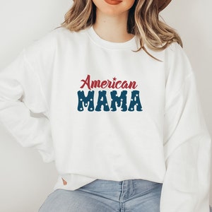 May include: White sweatshirt with the text "American MAMA" in red and blue. The word "American" is in a cursive font, and "MAMA" is in a block font with white stars. The sweatshirt is a casual style.