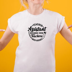 Pu&ograve; includere: Maglietta bianca con un grafico circolare nero che dice "Teacher Assistant Because Even Teachers Need Heroes" con un'icona di matita.