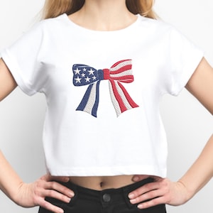 May include: White crop top featuring an embroidered bow design. The bow incorporates the colors of the American flag: blue with white stars, and red and white stripes. The shirt is a casual style.