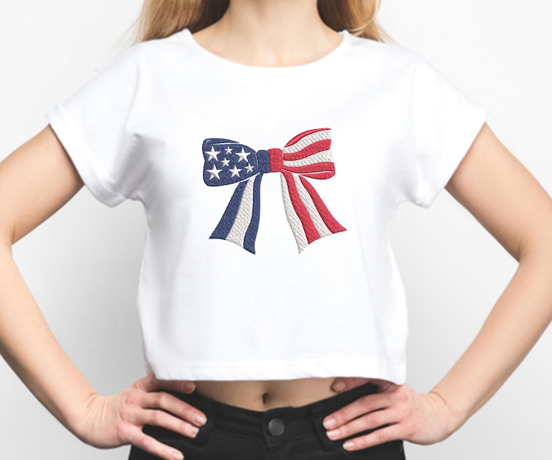 May include: White crop top featuring an embroidered bow design. The bow incorporates the colors of the American flag: blue with white stars, and red and white stripes. The shirt is a casual style.