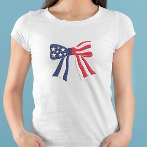 May include: White t-shirt featuring an embroidered bow design. The bow incorporates the colors of the American flag: blue with white stars, red and white stripes, and white accents. The shirt is a classic crew neck style.