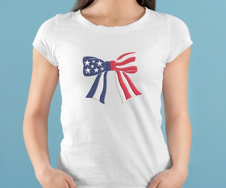 May include: White t-shirt featuring an embroidered bow design. The bow incorporates the colors of the American flag: blue with white stars, red and white stripes, and white accents. The shirt is a classic crew neck style.