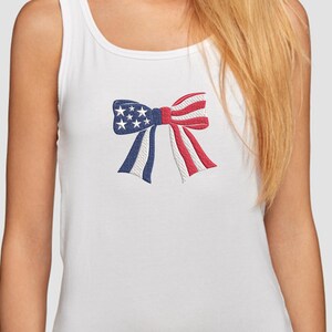 May include: White tank top with an embroidered bow design. The bow features a patriotic color scheme with blue sections adorned with white stars, and red and white striped sections, evoking an American flag theme.