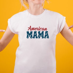 May include: White t-shirt with the text "American MAMA" in red and blue. The word "American" is in a script font, and "MAMA" is in a block font with star-shaped cutouts. The shirt is displayed on a person against a yellow background.