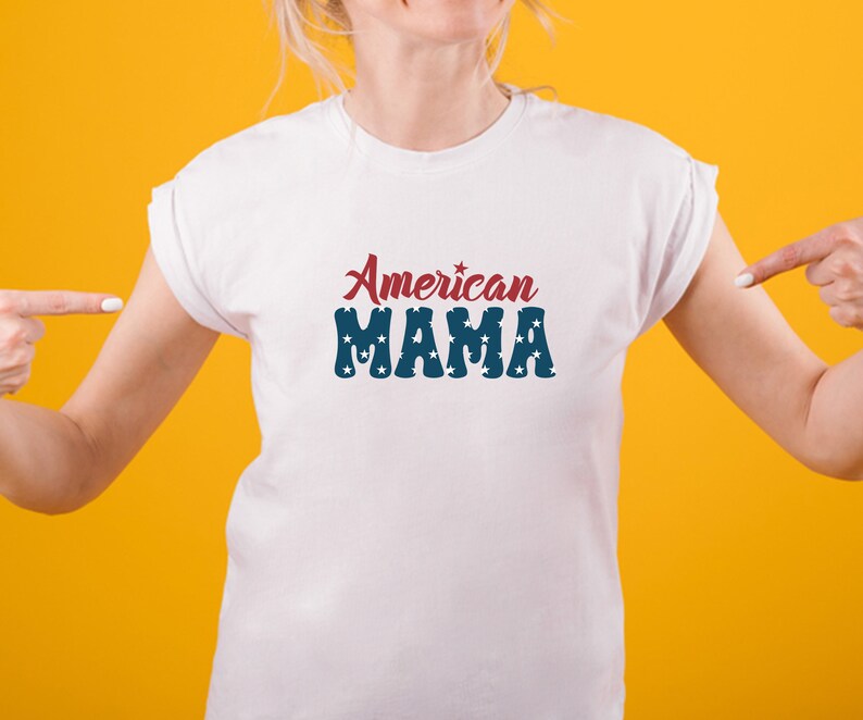 May include: White t-shirt with the text "American MAMA" in red and blue. The word "American" is in a script font, and "MAMA" is in a block font with star-shaped cutouts. The shirt is displayed on a person against a yellow background.