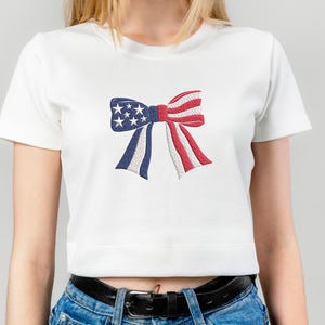 May include: White crop top featuring an embroidered bow design in red, white, and blue, with stars and stripes. The bow is a patriotic symbol, perfect for celebrating national holidays.
