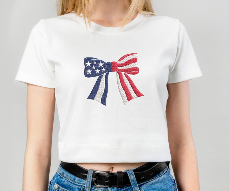 May include: White crop top featuring an embroidered bow design in red, white, and blue, with stars and stripes. The bow is a patriotic symbol, perfect for celebrating national holidays.
