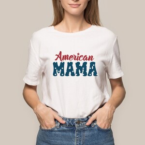 May include: White t-shirt with the text "American MAMA" in red and blue. The word "American" is in a script font, and "MAMA" is in a block font with star accents. The shirt is worn by a person in blue jeans.