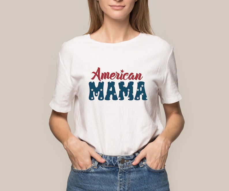 May include: White t-shirt with the text "American MAMA" in red and blue. The word "American" is in a script font, and "MAMA" is in a block font with star accents. The shirt is worn by a person in blue jeans.