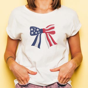 May include: White t-shirt featuring a patriotic bow design. The bow is embroidered with the colors of the American flag: blue with white stars, and red and white stripes. A casual and festive apparel item.