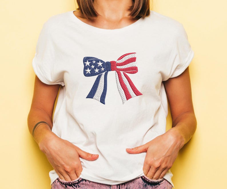 May include: White t-shirt featuring a patriotic bow design. The bow is embroidered with the colors of the American flag: blue with white stars, and red and white stripes. A casual and festive apparel item.