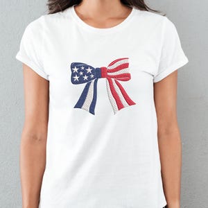 May include: White t-shirt featuring an embroidered bow design in the colors of the American flag. The bow has blue sections with white stars, red and white striped sections, and white rope-like accents. A patriotic apparel item.