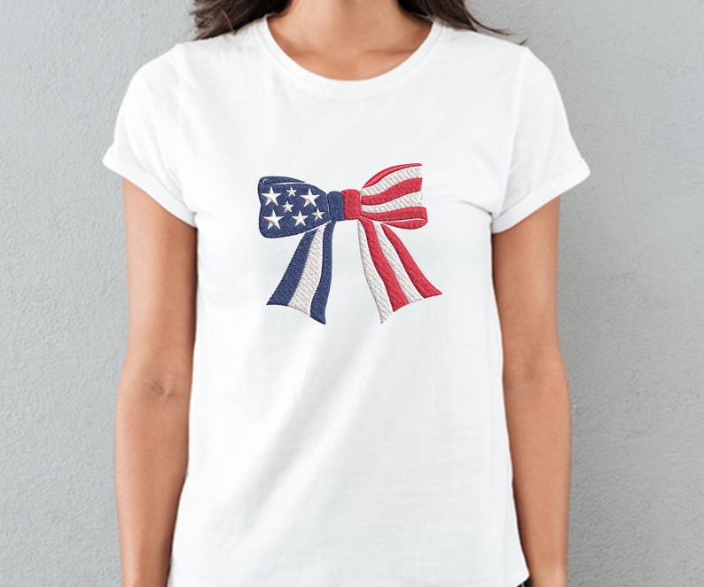 May include: White t-shirt featuring an embroidered bow design in the colors of the American flag. The bow has blue sections with white stars, red and white striped sections, and white rope-like accents. A patriotic apparel item.