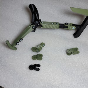 May include: A green and black 3D printed articulated robotic arm with multiple joints. The arm is disassembled and laid out on a white surface. The arm is made of plastic and has a carbon fibre pattern.